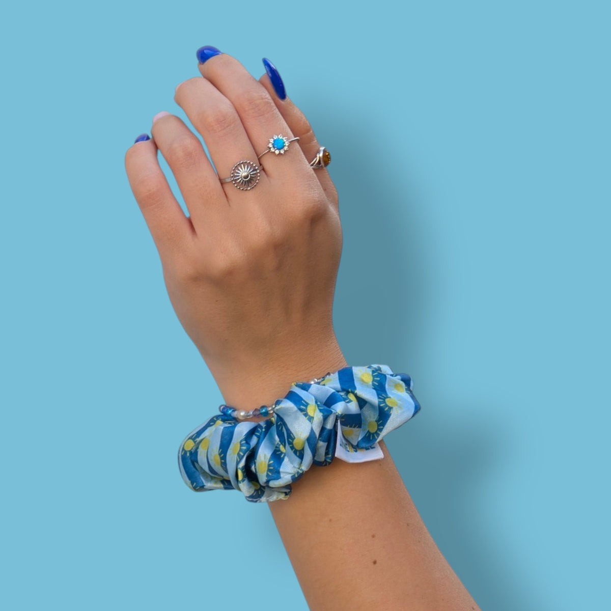 Chouchou Riviera Limone&Sole en satin bleu à rayures et motifs soleils porté au poignet d'une main manucurée