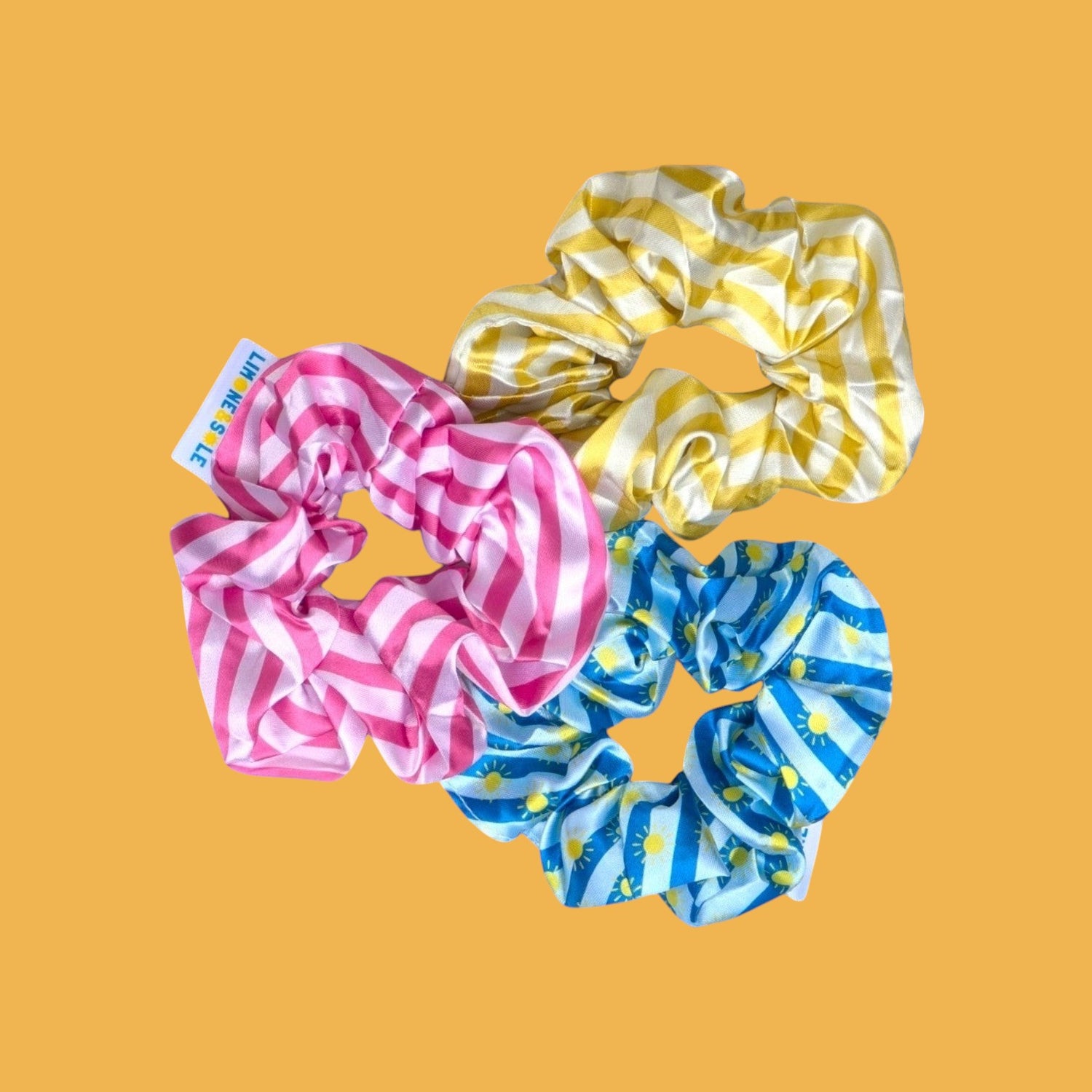 Trio de chouchous en satin Limone&Sole aux motifs rayés rose, jaune et bleu avec des soleils, texture brillante.