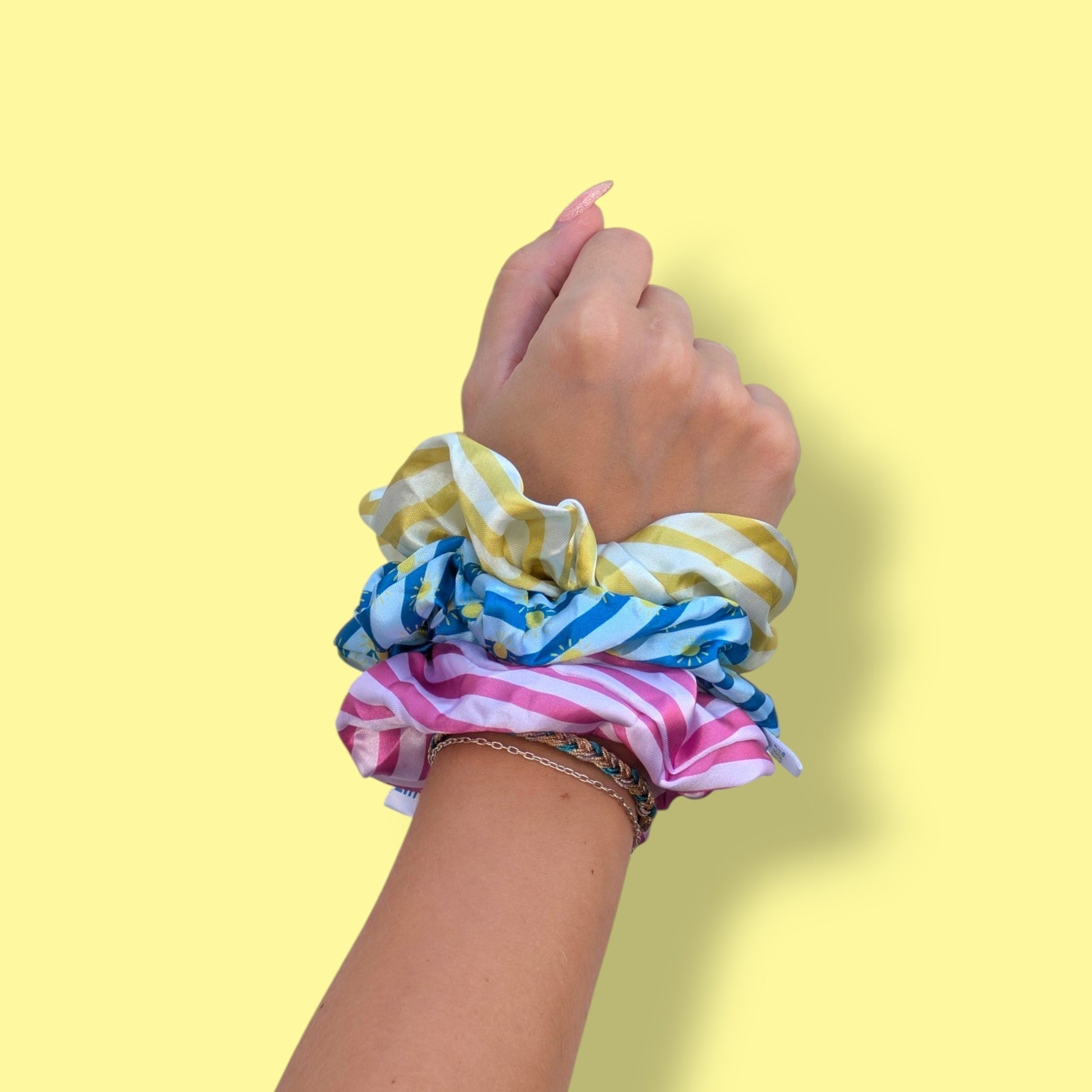 Trio de chouchous en satin Limone&Sole aux motifs rayés jaune, bleu et rose portés au poignet sur fond jaune